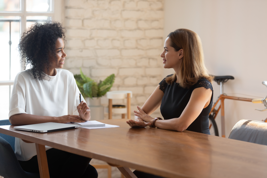Mulher em escritório conversando sobre promoção de cargo​ sentadas ao redor de uma mesa de reunião.